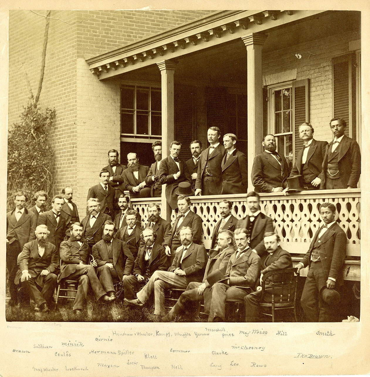 Photo of a group of men in suits, some on a porch and others in front of the porch
